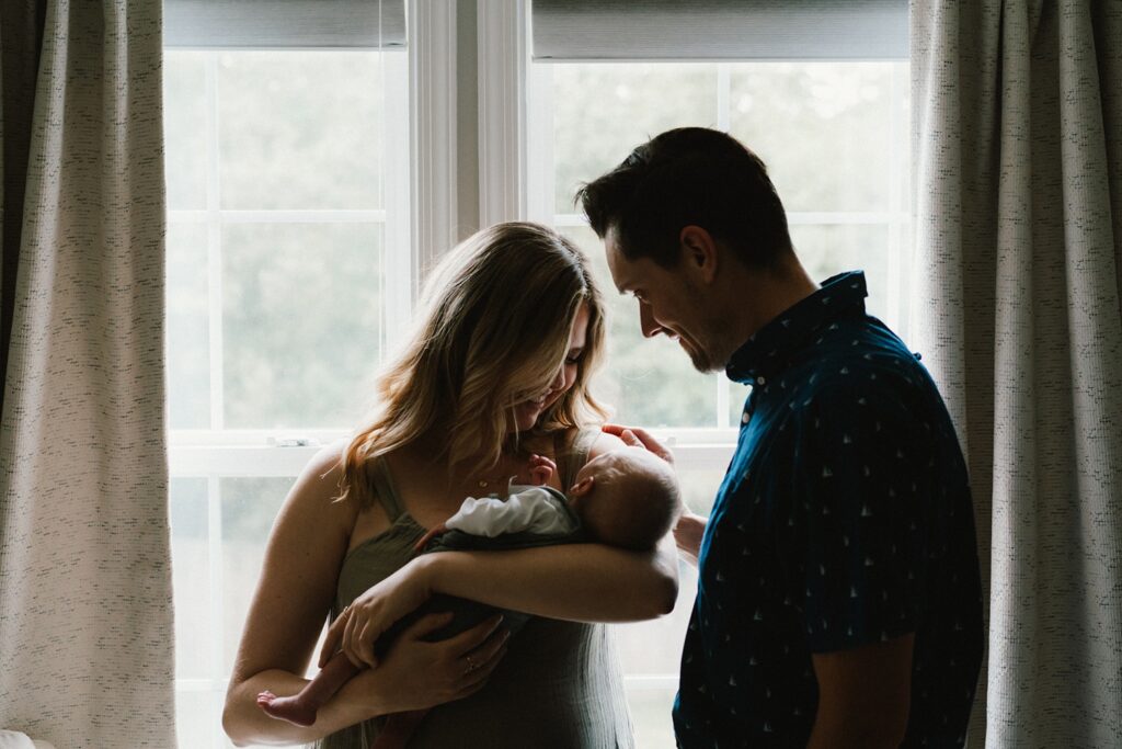 mom and dad with new baby boy in front of a window at home