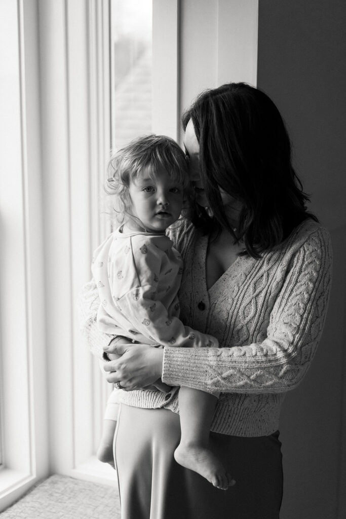 moody black and white photos of mother holding toddler by the window at home